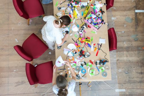 Zwei Kinder basteln an einem Tisch mit verschiedenen Bastelmaterialien wie Klebeband, Scheren, Papier und Farben.