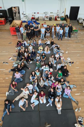 Eine große Gruppe junger Menschen sitzt und steht auf Matten in einer Halle und bildet die Form eines Menschen.