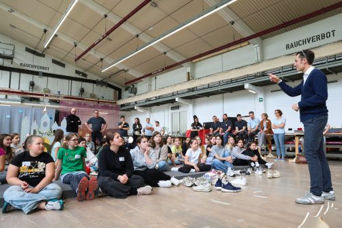 Ein Mann spricht vor einer Gruppe von sitzenden Kindern in einer großen Halle, während Erwachsene im Hintergrund zuschauen.