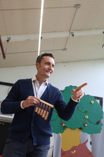 Ein lächelnder Mann mit einer Panflöte in der Hand zeigt mit dem Finger nach rechts, im Hintergrund steht ein grüner Pappbaum.