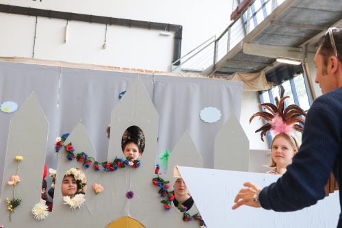 Mehrere Kinder blicken aus den Fenstern einer Burgkulisse, während ein Erwachsener mit ihnen spricht; einige Kinder tragen bunte Kostüme.