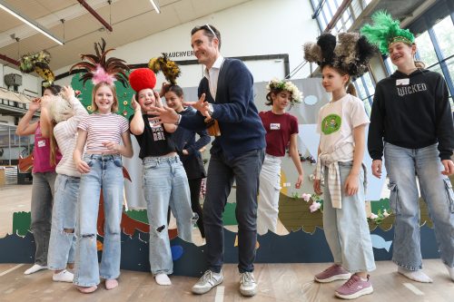 Eine Gruppe lachender Kinder mit bunten, auffälligen Kopfbedeckungen steht neben einem Erwachsenen in einem hellen Raum.