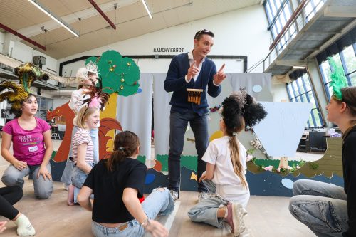 Ein Mann spielt inmitten von Kindern mit bunten Kostümen in einem hellen Raum mit großen Fenstern Theater.