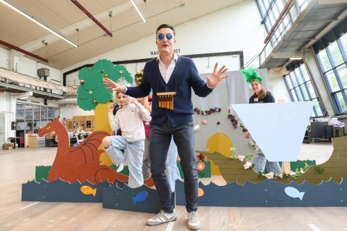 Ein Mann in blauem Pullover steht vor einer farbenfrohen Theaterkulisse mit zwei Kindern, die in einem Bastelprojekt mitwirken.