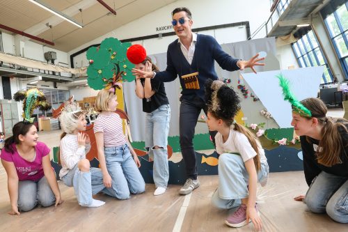 Ein Mann spielt mit fünf Kindern in bunten Kostümen vor einer Kulisse mit Bäumen und Fischen in einem hellen Raum Theater.
