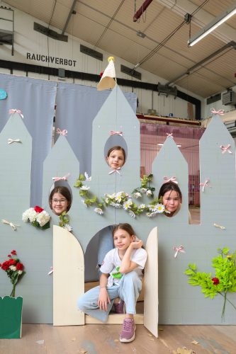 Vier Kinder posieren in einem selbstgebastelten Pappschloss, drei schauen durch Fenster, eines sitzt in der Türöffnung davor.