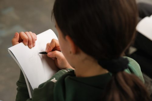 Ein Kind mit dunklen Haaren zeichnet mit einem schwarzen Stift auf ein weißes Blatt Papier in einem Skizzenbuch.