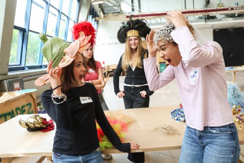 Vier junge Frauen in einem hellen Raum lachen und probieren bunte, auffällige Hüte an.