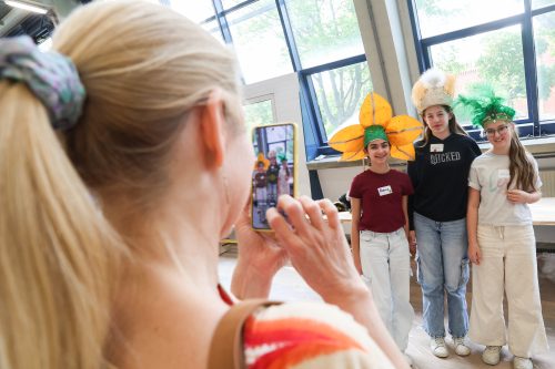 Eine Frau fotografiert drei Kinder, die bunte und fantasievolle Hüte tragen, in einem hellen Raum mit großen Fenstern.