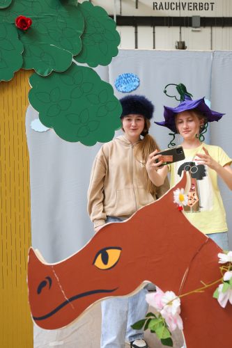 Zwei Kinder stehen hinter einer Papp-Dinosaurierfigur und einem Pappbaum; beide tragen kreative Hüte und machen ein Selfie.