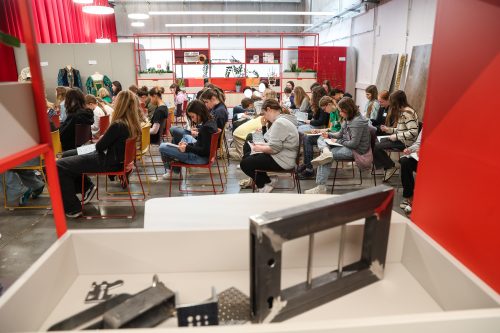 Viele junge Menschen sitzen in einem hellen Raum auf Stühlen und schreiben konzentriert auf Papier.