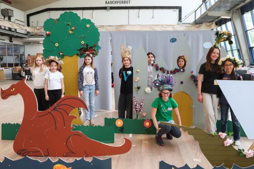 Acht Kinder stehen in einer bunt dekorierten Kulisse mit einem roten Drachen, Bäumen und Blumen, einige tragen Kostüme und Kopfschmuck.