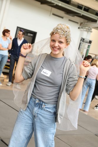 Junge Frau mit Namensschild „Annalena“, trägt einen glitzernden Kopfschmuck und einen Schleier, lächelt und steht in einer Halle.