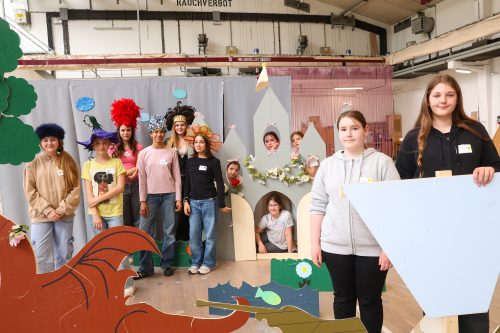 Eine Gruppe von Kindern in bunten Kostümen steht und sitzt vor einer märchenhaften Kulisse in einem großen Innenraum.