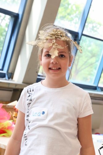 Ein Mädchen mit einem goldenen Kopfschmuck und einem Namensschild „Lea“ lächelt in einem hellen Raum mit großen Fenstern.