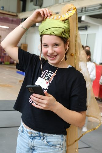 Ein lächelndes Mädchen mit einem grünen Turban und einem goldenen Schleier hält ein Smartphone in der Hand.