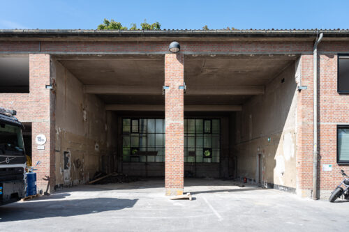 Offene Backstein-Garagenhalle mit großen Glastüren im Hintergrund und sichtbaren Gebrauchsspuren an den Wänden.