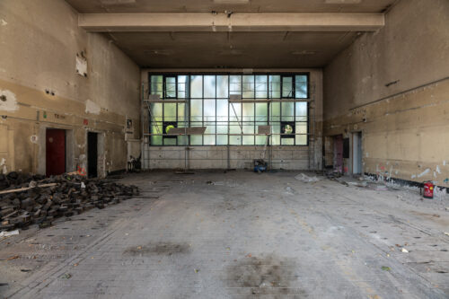 Leere, heruntergekommene Industriehalle mit großen, teils beschädigten Fenstern und verstreuten Trümmern auf dem Boden.