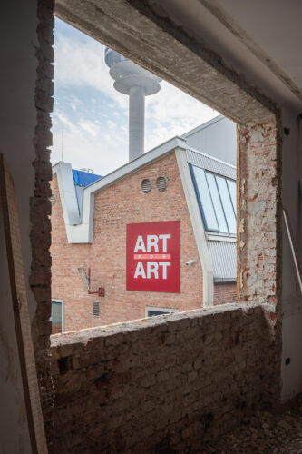 Blick durch eine große, rechteckige Bauöffnung auf ein Backsteingebäude mit rotem Schild „ART FOR ART“.