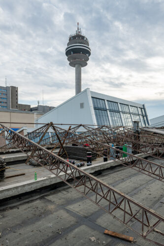 Eine Baustelle mit Stahlträgern im Vordergrund, dahinter ein moderner Turm und ein Gebäude mit schrägem Glasdach.