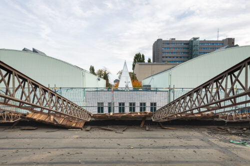 Eine teilweise abgebaute Eisenbrücke mit Bauzaun, dahinter moderne Gebäude und ein spitzes weißes Bauwerk.