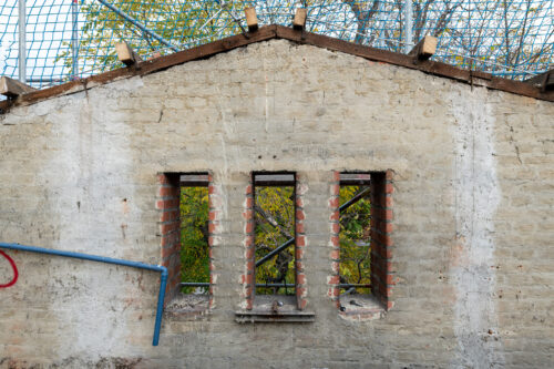 Alte Backsteinmauer mit drei schmalen Fenstern, durch die Bewuchs und ein Zaun im Hintergrund sichtbar sind.