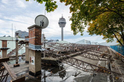 Ein eingestürztes Metalldach auf einem Gebäudedach mit Pfützen, daneben Bäume und im Hintergrund ein hoher Fernsehturm.