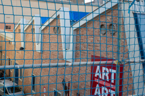 Backsteingebäude mit Lüftungsschlitzen und einem roten Schild mit dem Wort „ART“, von einem blauen Netz teilweise verdeckt.