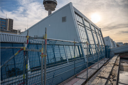 Gebäude mit schrägen Glasfassaden und Baugerüst im Vordergrund, im Hintergrund ein hoher Turm bei Sonnenuntergang.