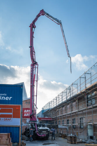 Ein großer Betonpumpenwagen mit auffällig langem Ausleger steht auf einer Baustelle zwischen Gebäuden.