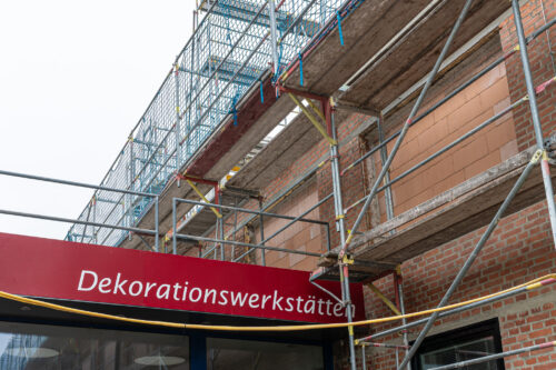 Ein rotes Schild mit der Aufschrift „Dekorationswerkstätten“ hängt an einem Gebäude, das mit Gerüsten umgeben ist.