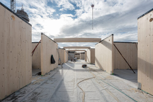Eine Baustelle mit Holzwänden und offenen Decken, Kabel und Bauwerkzeuge liegen am Boden, der Himmel ist teilweise bewölkt.