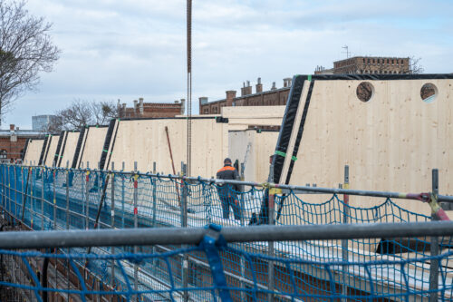 Mehrere Holzhäuser werden auf einer Baustelle errichtet, während Arbeiter auf dem Gerüst arbeiten.