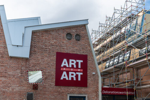 Backsteinfassade mit großem roten Schild "ART FOR ART", daneben ein Gerüst und ein Eingang mit dem Schriftzug "Dekorationswerkstätten".