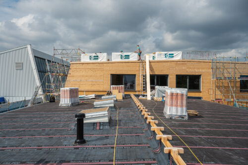 Flachdach einer Baustelle mit verlegter Abdichtungsfolie, Baumaterialien und mehreren Arbeitern auf dem Dach eines Holzgebäudes.