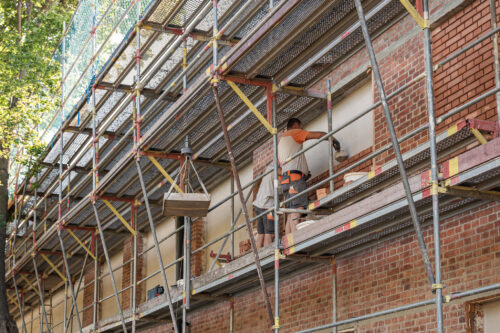 Zwei Bauarbeiter stehen auf einem Gerüst und arbeiten an der Fassade eines roten Backsteingebäudes.