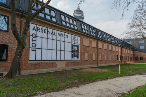 Backstein-Gebäude mit großem Schild „Foto Arsenal Wien“ an der Wand, davor eine Wiese und ein Gehweg, Fernsehturm im Hintergrund.