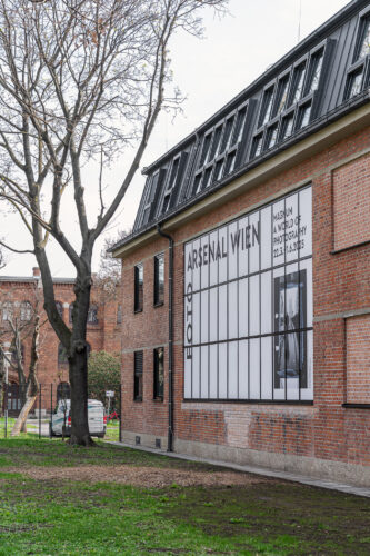 Backsteingebäude mit einem großen Plakat, das „ARSENAL WIEN“ und Informationen zu einer Fotoausstellung zeigt.