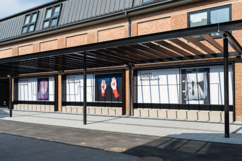 Backstein-Gebäude mit großen Fenstern und Werbeplakaten für "Magnum World of Photography" an der Außenwand.