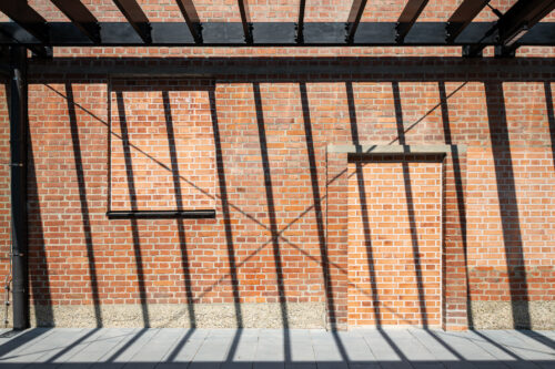 Backsteinwand mit rechteckigem Fenster und Tür, über die sich die Schatten von Stahlträgern diagonal ziehen.