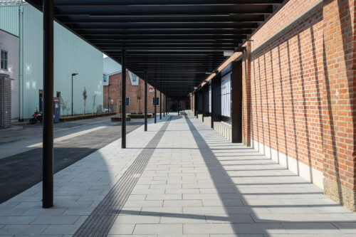 Überdachter Gehweg mit Schattenmustern neben einer Backsteinmauer in einem modernen städtischen Bereich.