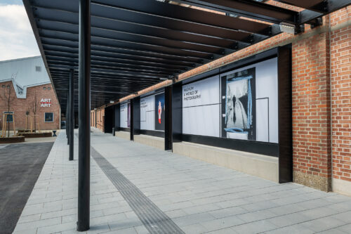 Überdachter Gehweg entlang einer Backsteinmauer mit großen Plakaten, die Fotografiekunst und den Text „A World of Photography“ zeigen.