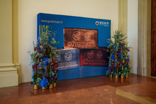 Drei große, dekorative Ziegel mit der Aufschrift "Güteziegel" stehen vor einem blauen Hintergrund, umrahmt von Blumenarrangements.