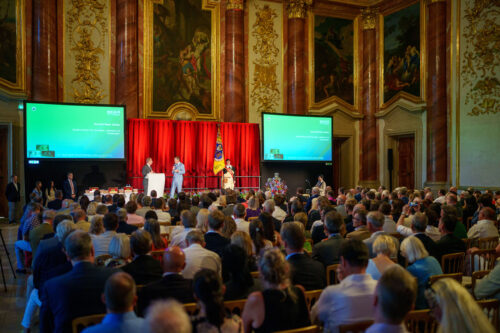Großer festlicher Saal mit vielen sitzenden Menschen, einer Bühne, Rednern am Mikrofon und zwei großen Projektionsleinwänden.