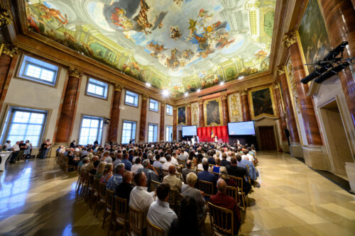Viele Menschen sitzen in einem prächtigen Saal mit bemalter Decke und verfolgen eine Präsentation auf zwei Bildschirmen vorne.