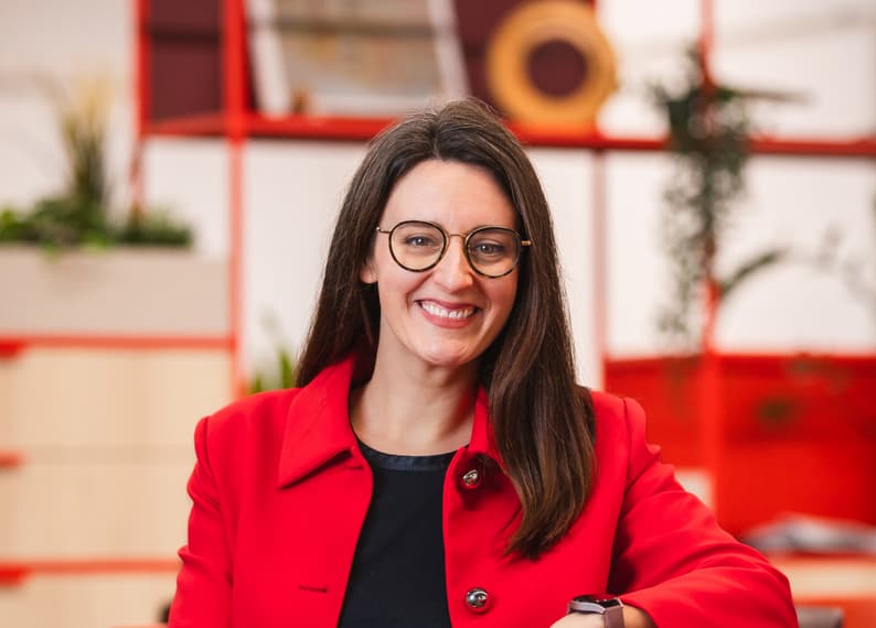 Frau mit langen braunen Haaren und Brille lächelt, trägt einen roten Blazer und sitzt vor einem modernen Regal im Hintergrund.