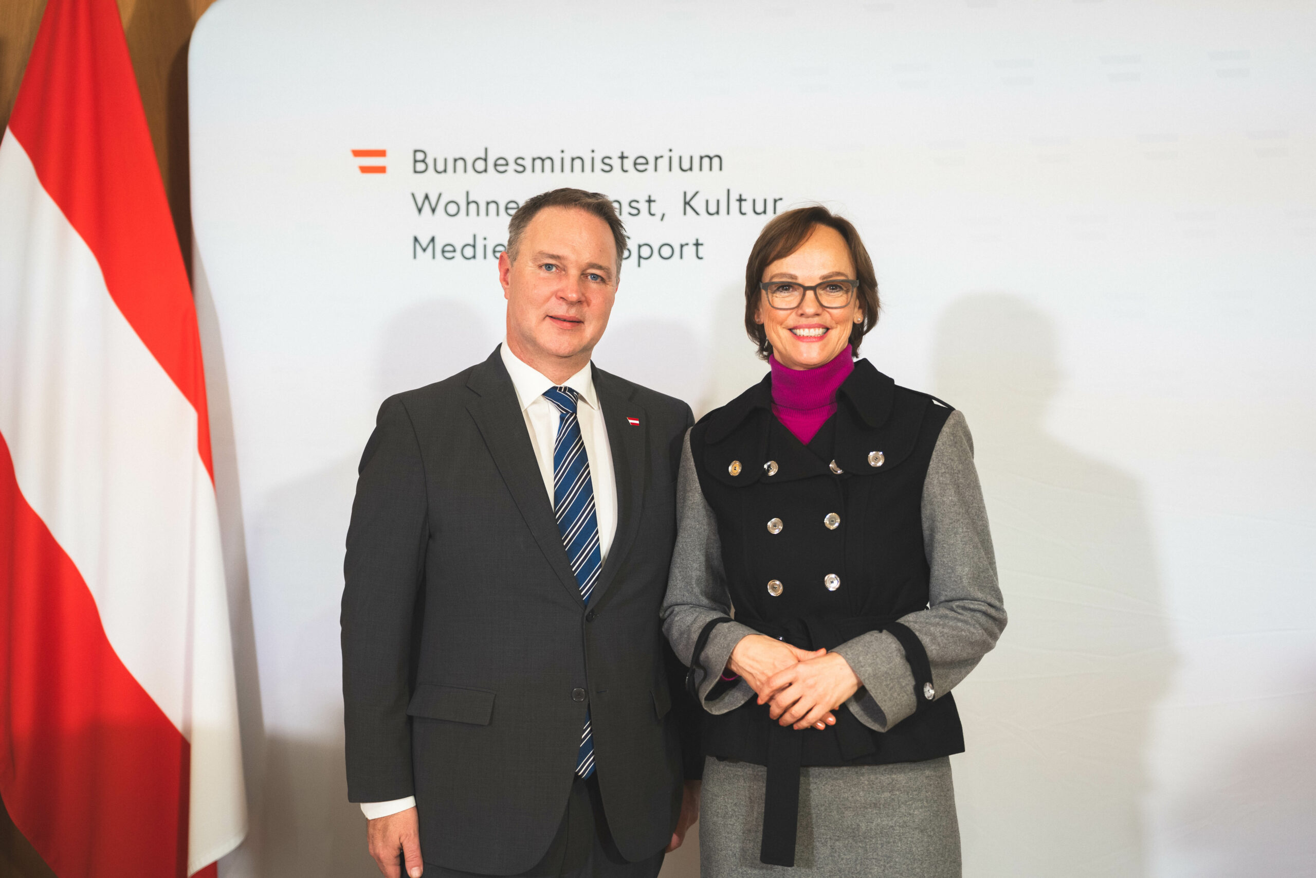 Ein Mann und eine Frau stehen nebeneinander vor einer Wand mit dem Logo des Bundesministeriums für Kunst, Kultur, Medien und Sport.