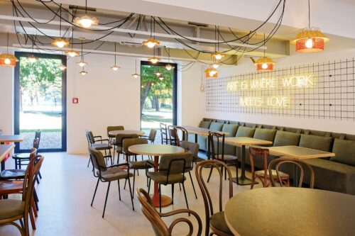 Modernes Café mit runden Tischen, verschiedenen Stühlen, Polsterbank und Leuchtschrift „ART IS WHERE WORK MEETS LOVE“ an der Wand.