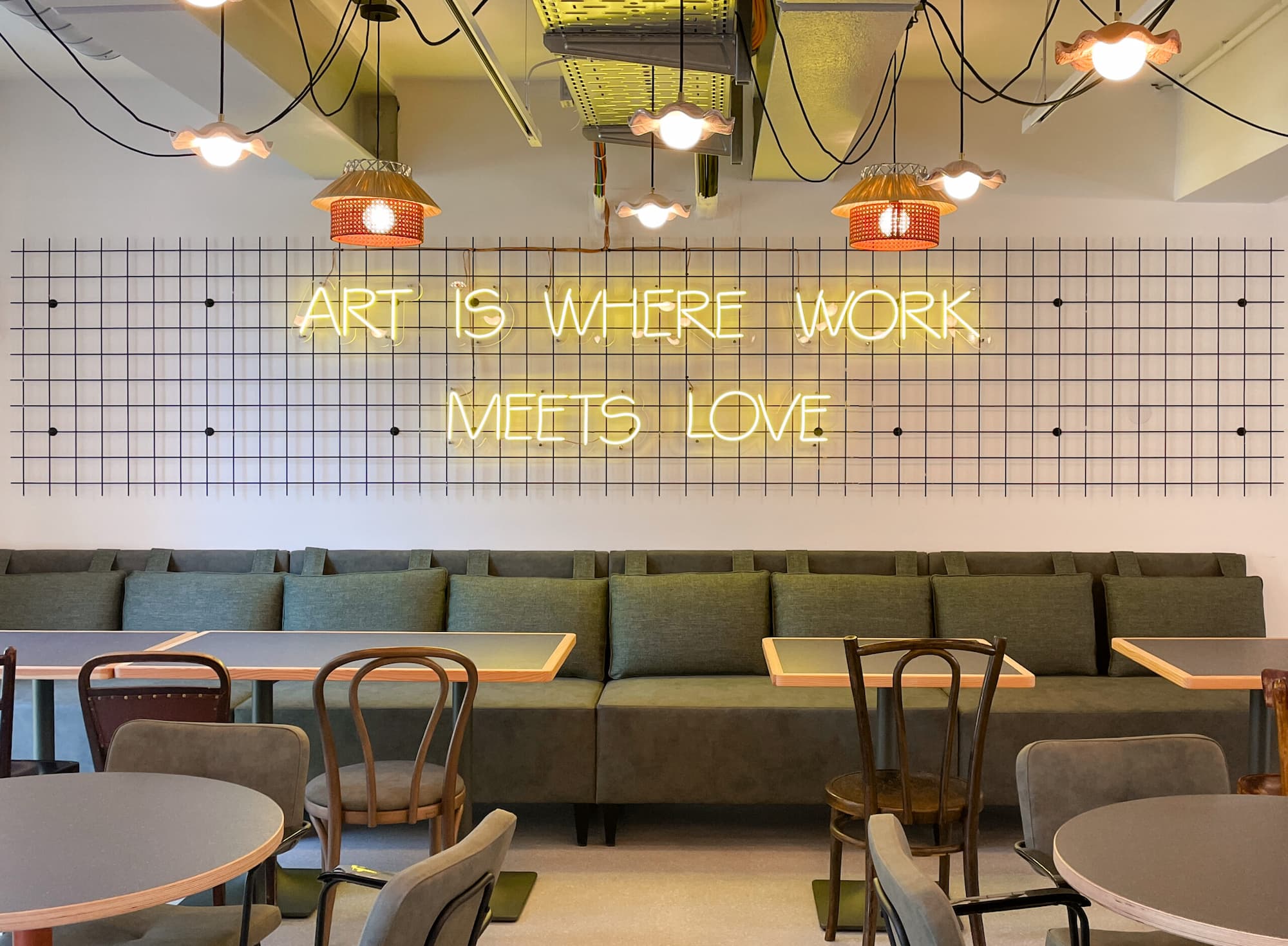 Industriell eingerichtetes Café mit grüner Sitzbank und gelber Leuchtschrift an der Wand: „ART IS WHERE WORK MEETS LOVE“.