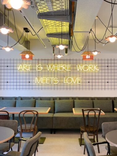 Moderne Café- oder Loungebereich mit grüner Sitzbank, Holzmöbeln und einem Leuchtschild mit der Aufschrift „ART IS WHERE WORK MEETS LOVE“.